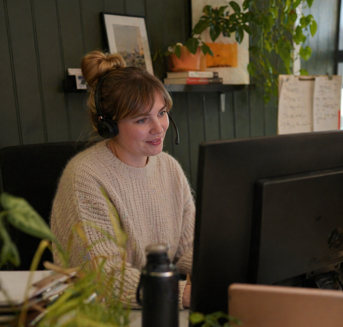 Person wearing a headset sitting at a computer in an office setting with plants and decor.